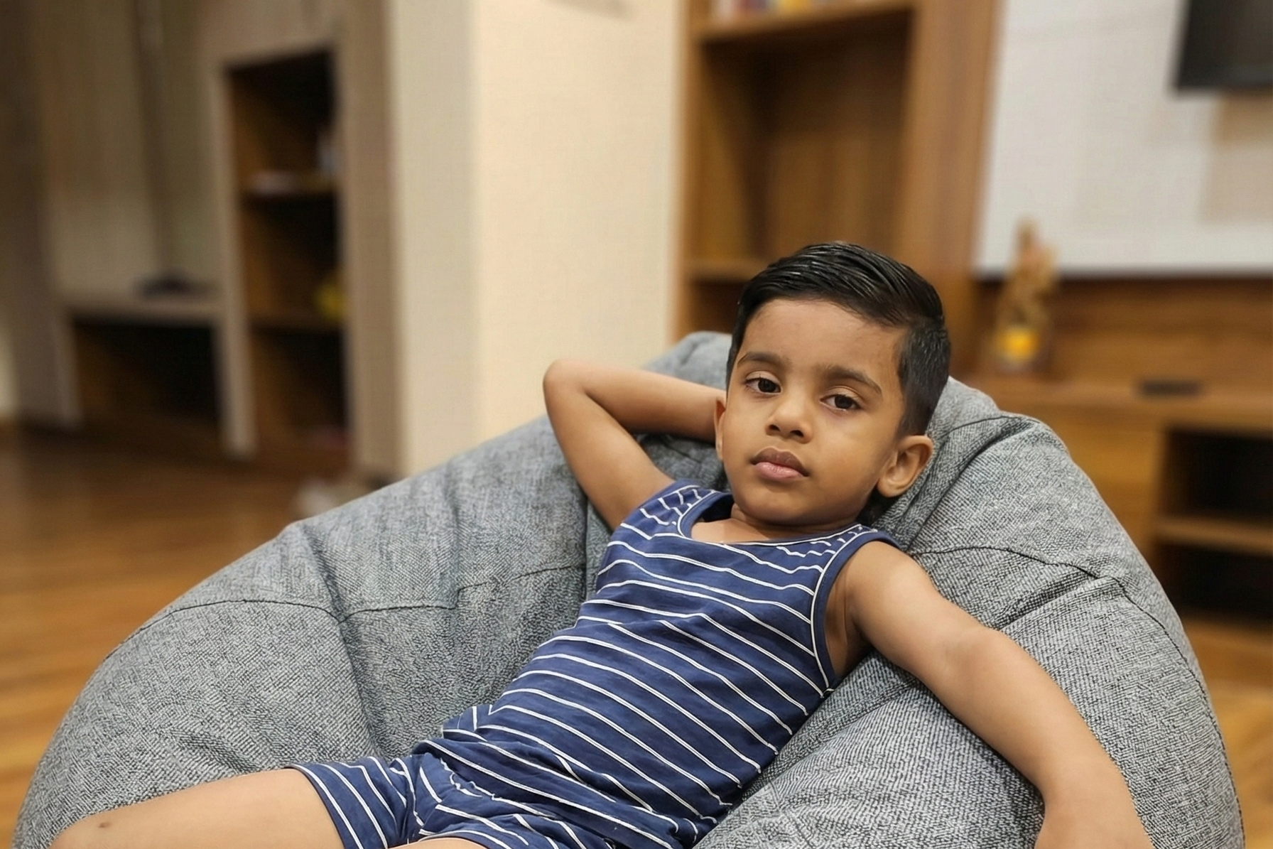 Child sitting on a gray bean bag chair in a room with wooden flooring and furniture.